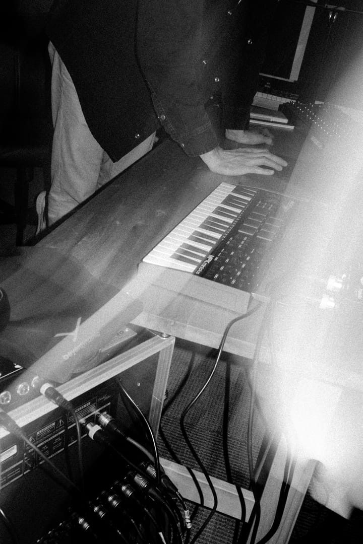 Hands on a keyboard above a rack of synthesizers, black and white